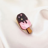 A pink and chocolate handcrafted popsicle pin adorned with three sparkling rhinestones rests on a soft, silky white fabric background.