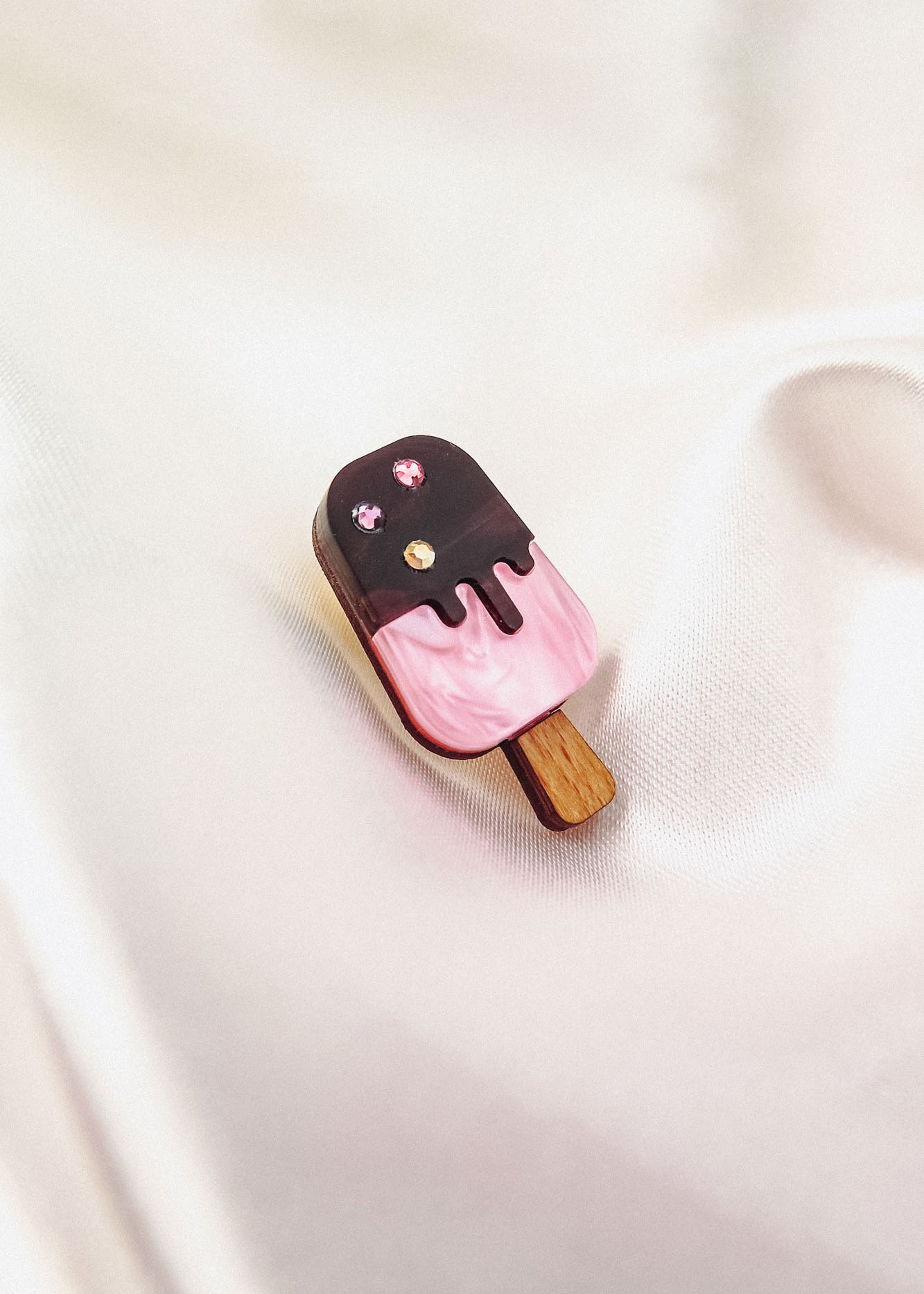 A pink and chocolate handcrafted popsicle pin adorned with three sparkling rhinestones rests on a soft, silky white fabric background.