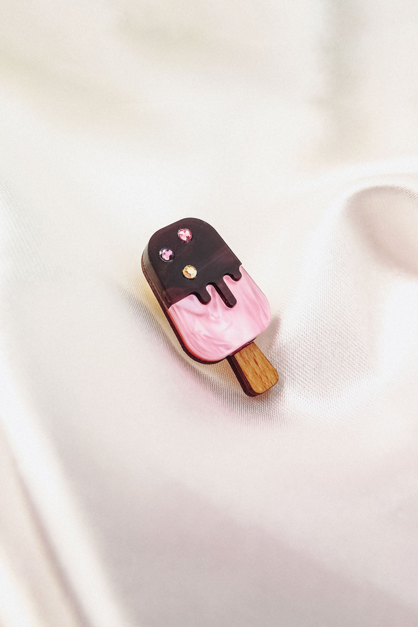 A pink and chocolate handcrafted popsicle pin adorned with three sparkling rhinestones rests on a soft, silky white fabric background.