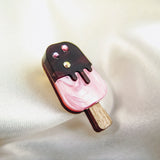 A handcrafted popsicle pin shaped like a pink ice cream bar with a chocolate coating, topped with three small rhinestones, lies on a soft, cream-colored fabric background.