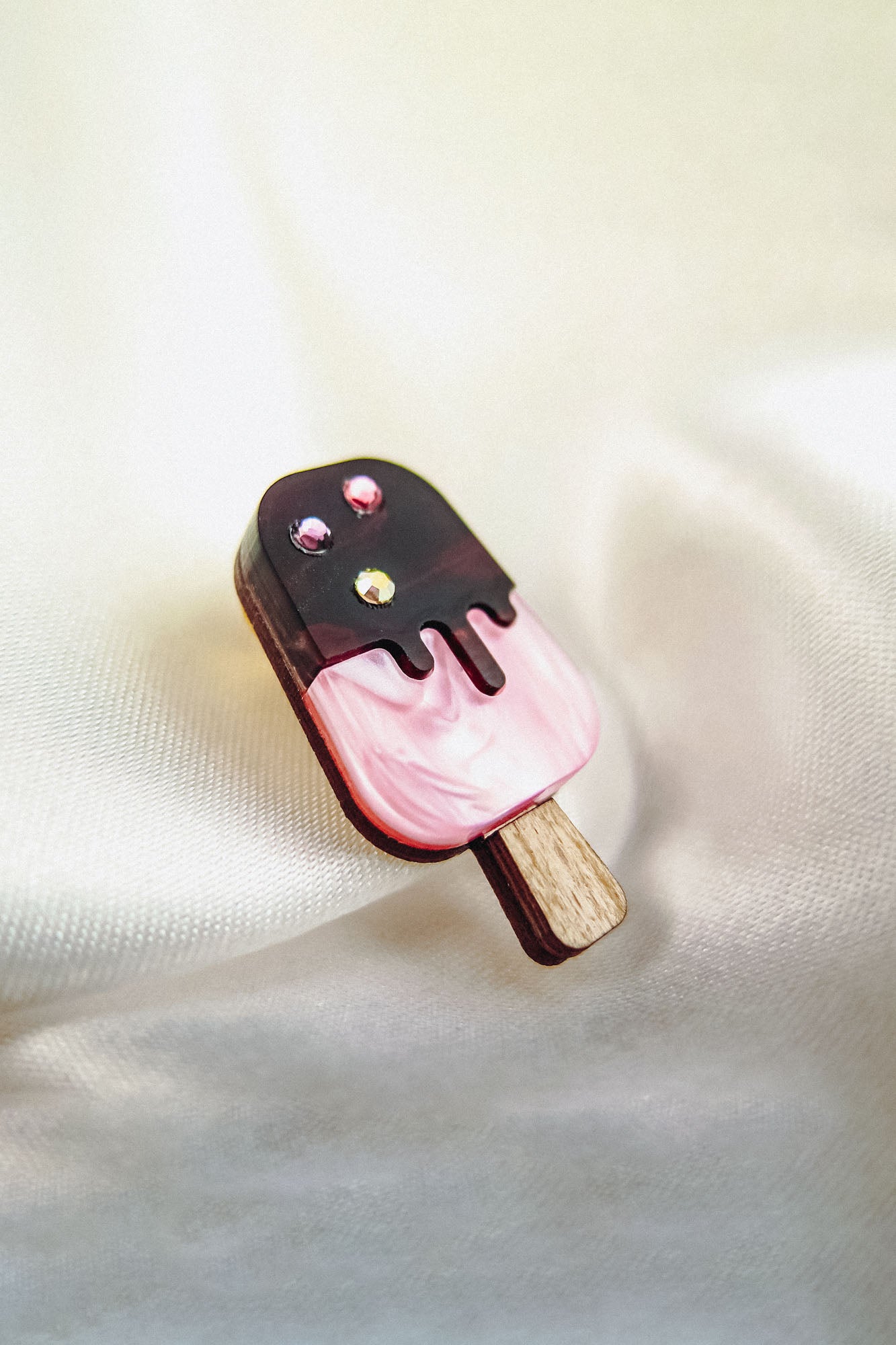 A handcrafted popsicle pin shaped like a pink ice cream bar with a chocolate coating, topped with three small rhinestones, lies on a soft, cream-colored fabric background.