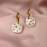 A pair of lightweight hoop earrings with irregular white pendants, each adorned with small colorful gems, rest on soft, pink satin fabric—handcrafted earrings perfect for adding sparkle to any look.