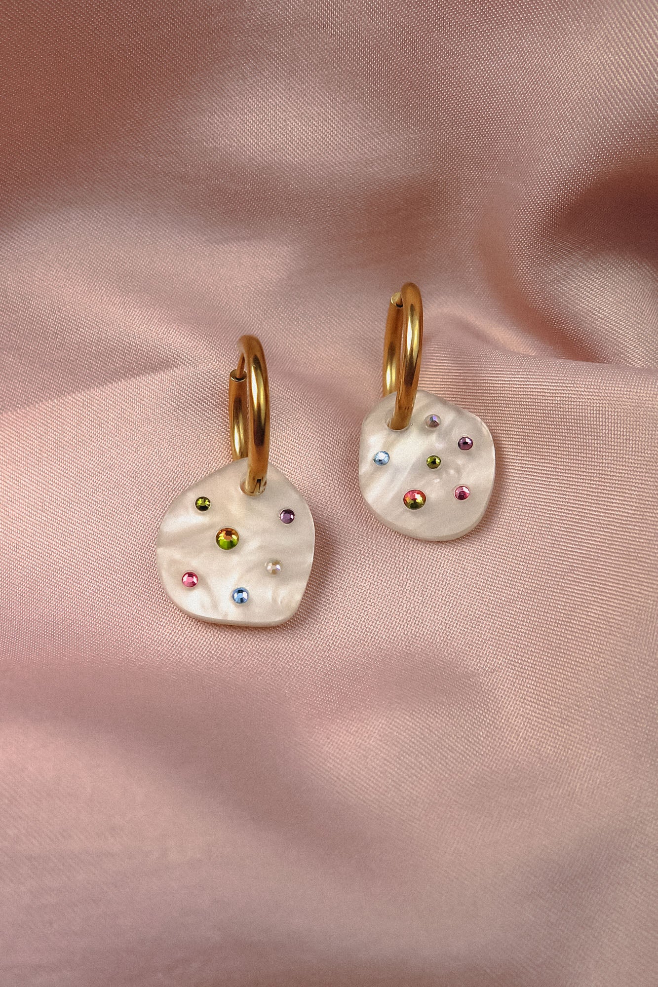 A pair of lightweight hoop earrings with irregular white pendants, each adorned with small colorful gems, rest on soft, pink satin fabric—handcrafted earrings perfect for adding sparkle to any look.