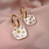 Lightweight hoop earrings with irregular white pearl-like drops, each decorated with colorful small gemstones, displayed on soft pink satin fabric.