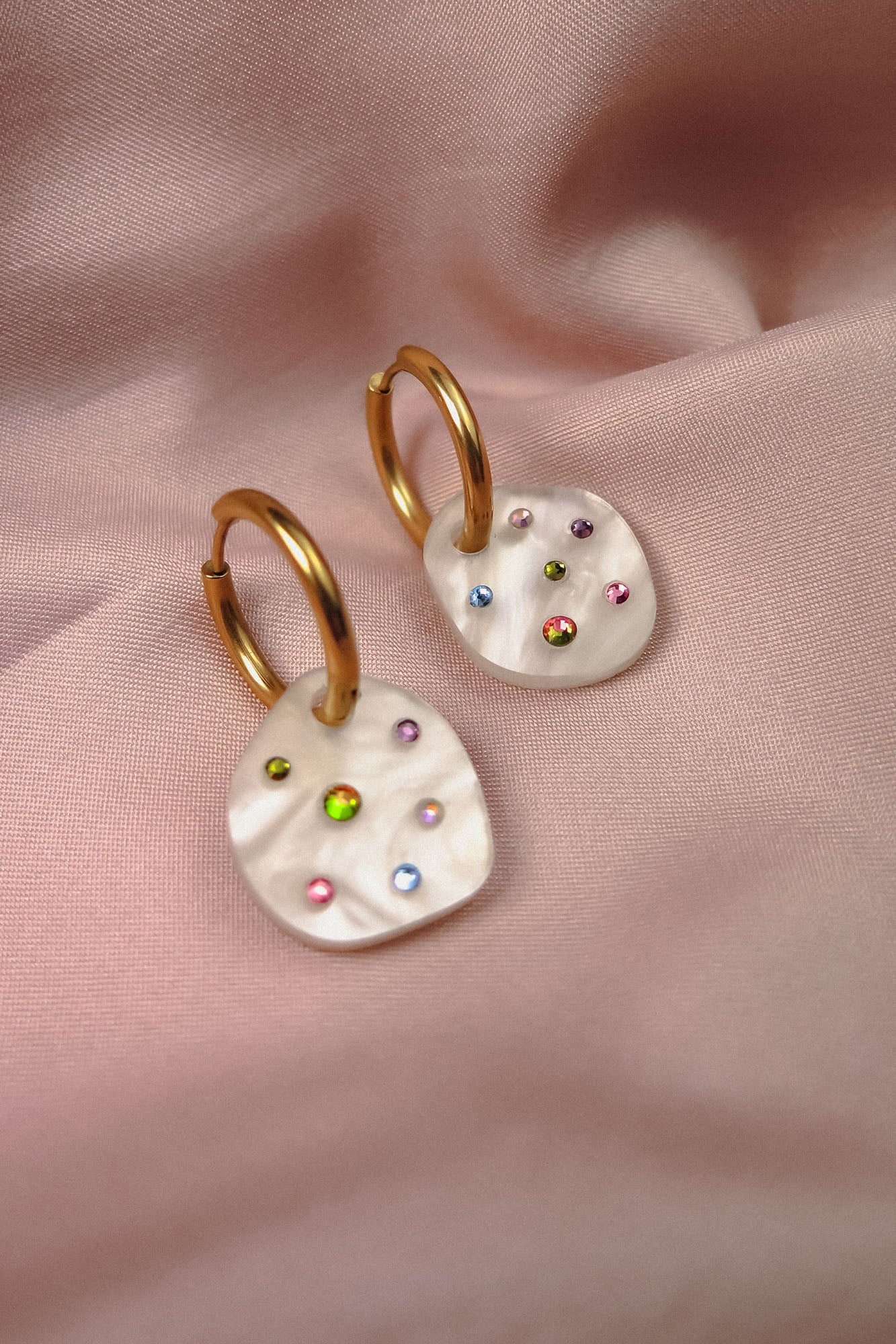 Lightweight hoop earrings with irregular white pearl-like drops, each decorated with colorful small gemstones, displayed on soft pink satin fabric.