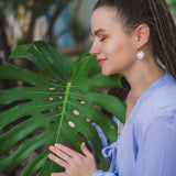 A woman with long hair in a ponytail, wearing a light blue top and lightweight hoop earrings, gently touches a large green monstera leaf while smiling and closing her eyes outdoors.