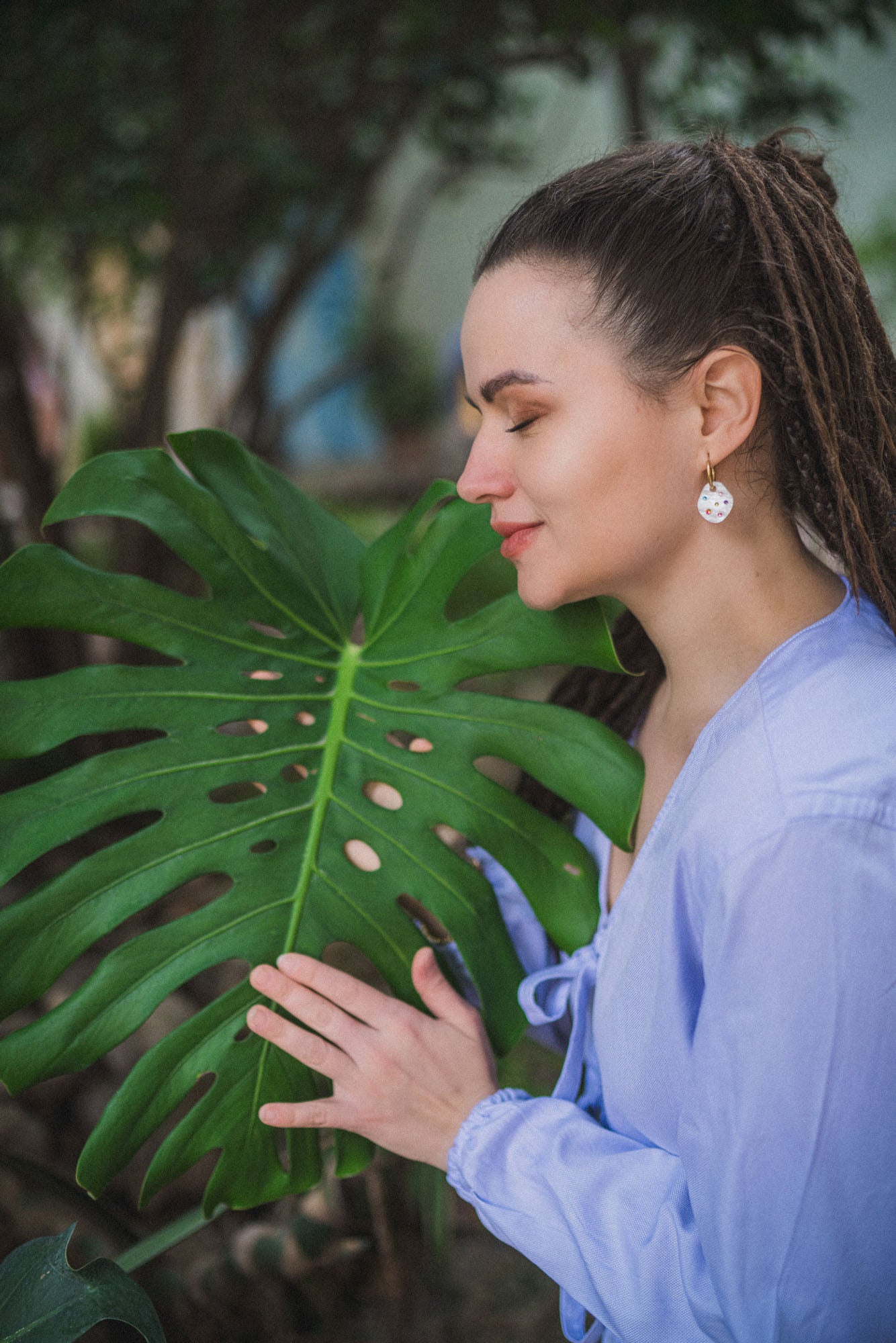 A woman with long hair in a ponytail, wearing a light blue top and lightweight hoop earrings, gently touches a large green monstera leaf while smiling and closing her eyes outdoors.