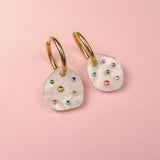 A pair of lightweight hoop earrings with irregular white, shell-like pendants, each adorned with small, colorful gemstone studs, displayed on a light pink background.