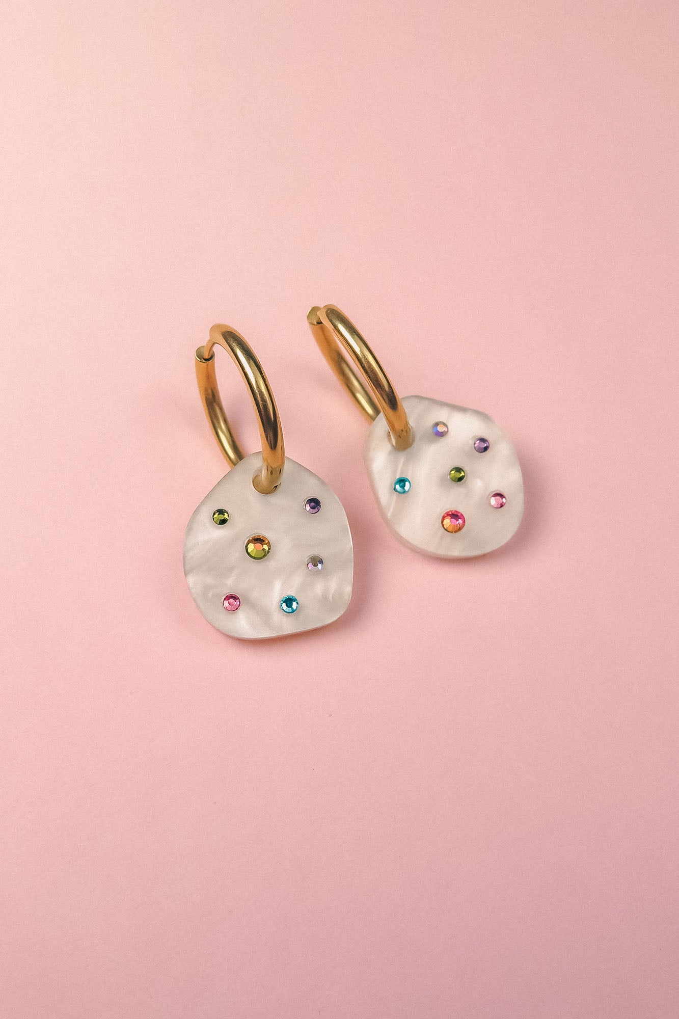 A pair of lightweight hoop earrings with irregular white, shell-like pendants, each adorned with small, colorful gemstone studs, displayed on a light pink background.