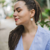 A woman with long, brown dreadlocks and rainbow rhinestone earrings stands outdoors with her eyes closed, wearing a light blue blouse, appearing calm and peaceful.