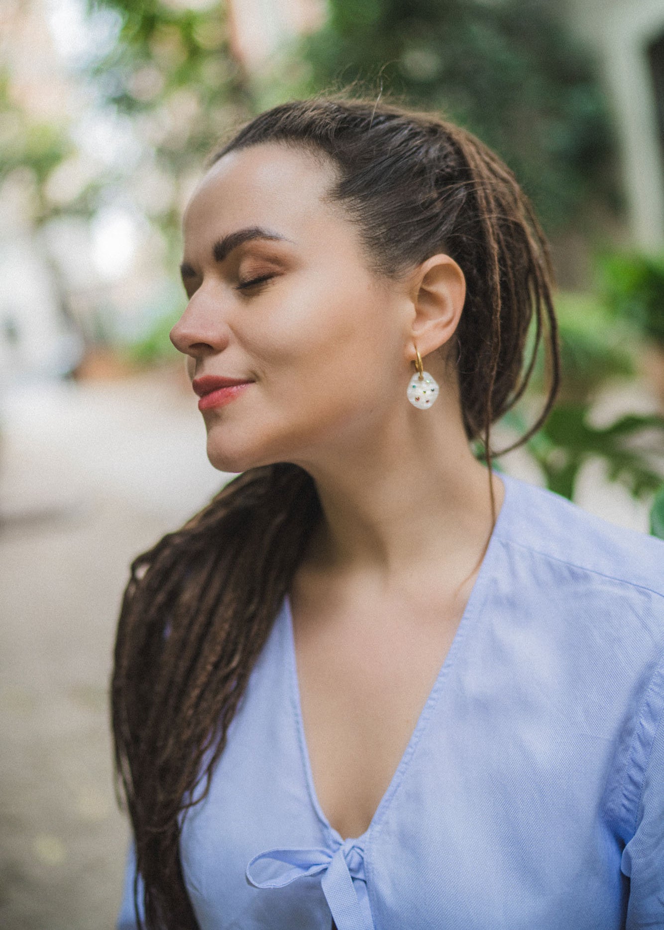 A woman with long, brown dreadlocks and rainbow rhinestone earrings stands outdoors with her eyes closed, wearing a light blue blouse, appearing calm and peaceful.