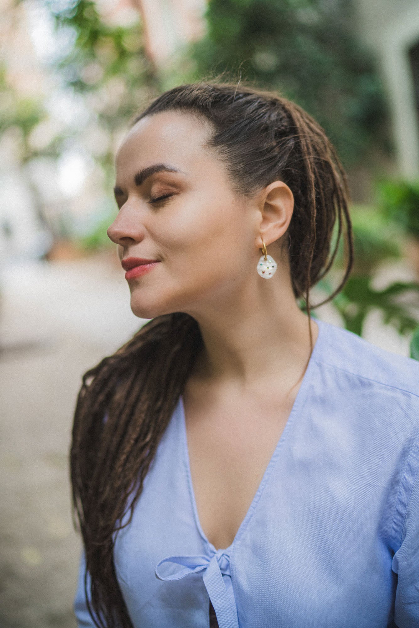 A woman with long, brown dreadlocks and rainbow rhinestone earrings stands outdoors with her eyes closed, wearing a light blue blouse, appearing calm and peaceful.