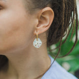 Close-up of a person with brown hair in dreadlocks wearing handcrafted, translucent round earrings with small colorful beads and a light blue top, set against a green leafy background.