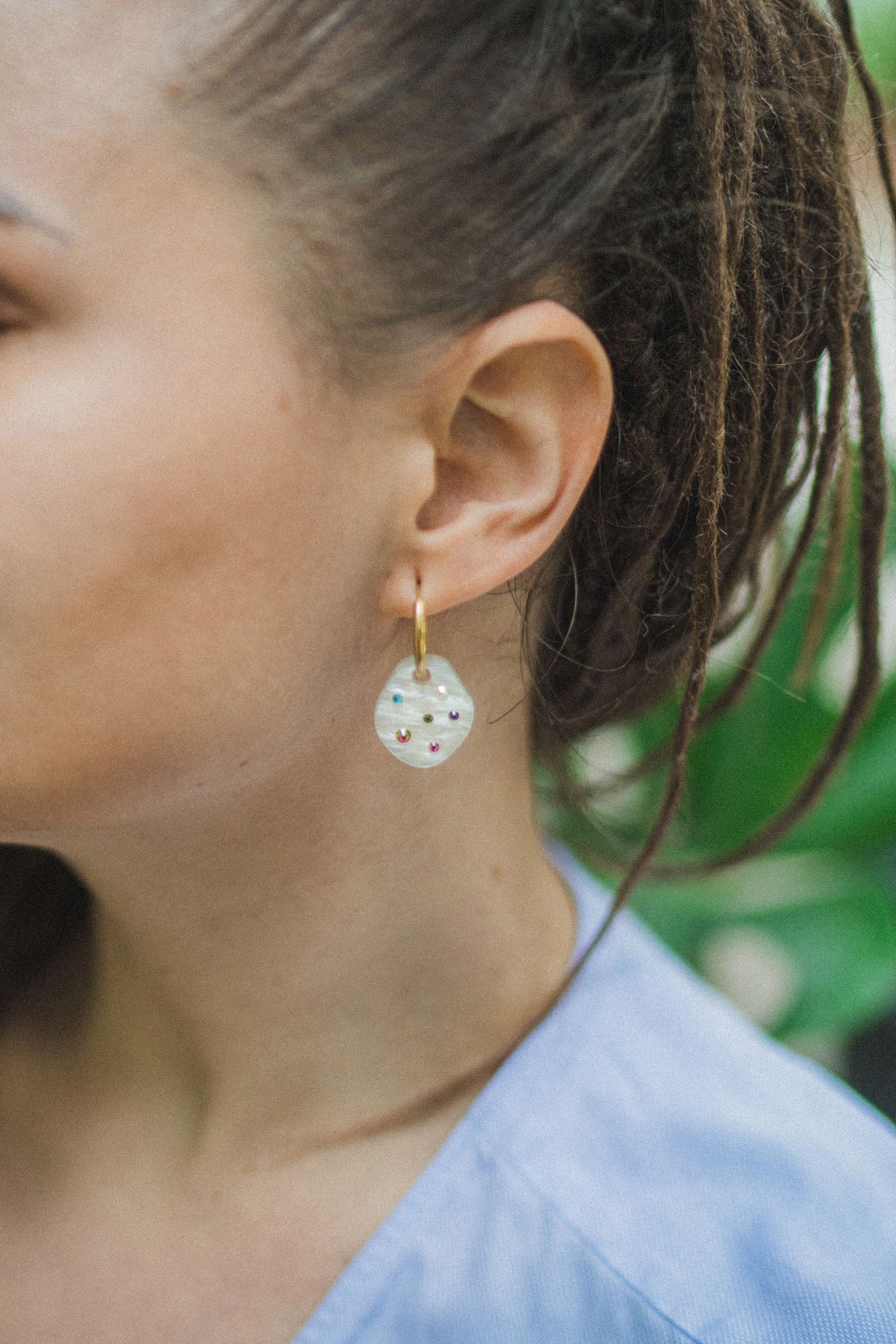 Close-up of a person with brown hair in dreadlocks wearing handcrafted, translucent round earrings with small colorful beads and a light blue top, set against a green leafy background.