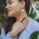 A woman with long braided hair wearing a light blue top smiles with her eyes closed, gently touching her neck and displaying a ring and handcrafted earrings, standing near green leaves.