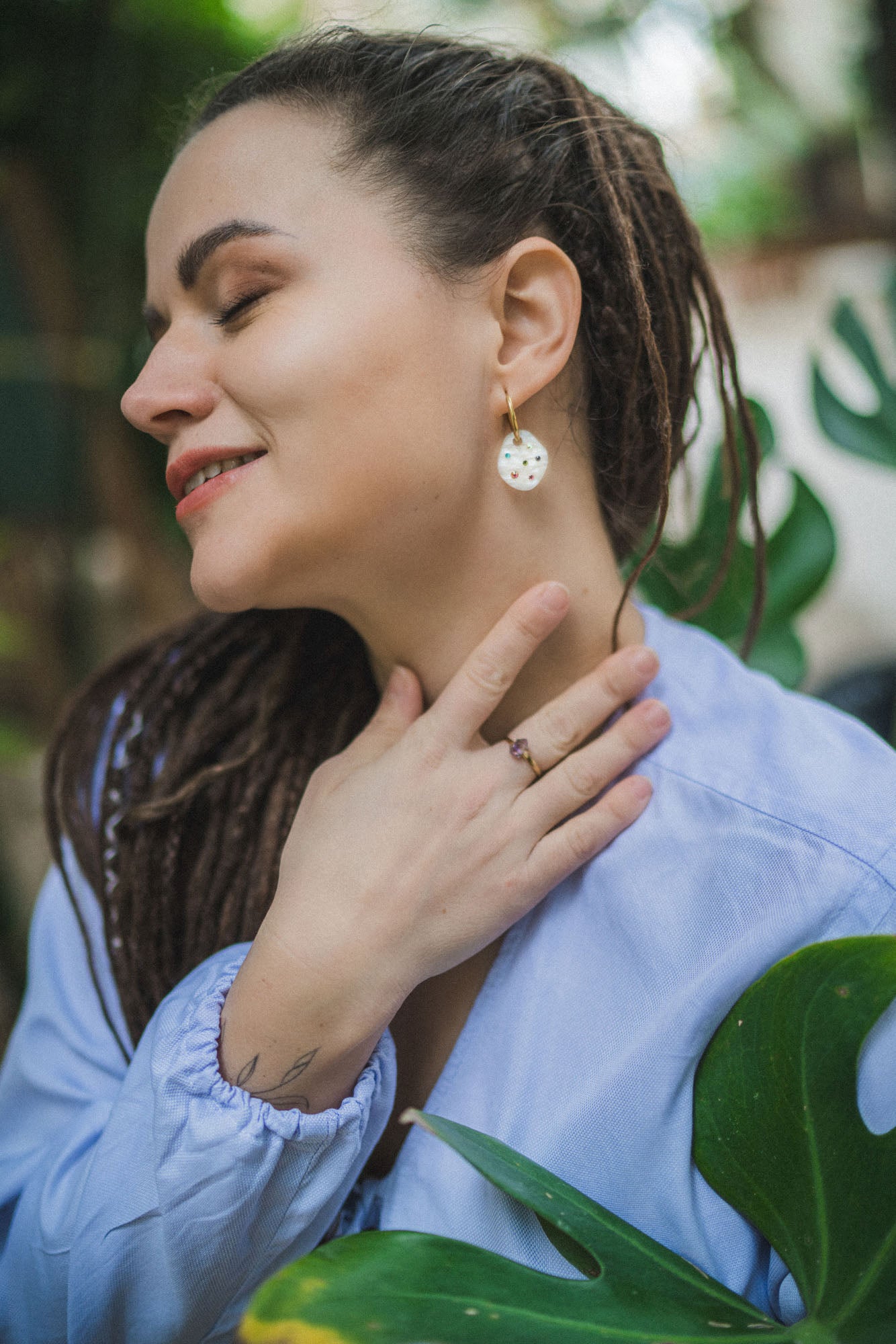 A woman with long braided hair wearing a light blue top smiles with her eyes closed, gently touching her neck and displaying a ring and handcrafted earrings, standing near green leaves.