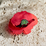 A red brooch clip shaped like a flower with a green acrylic glass center rests on a textured, off-white stone surface, accented by delicate dried plants in the background.