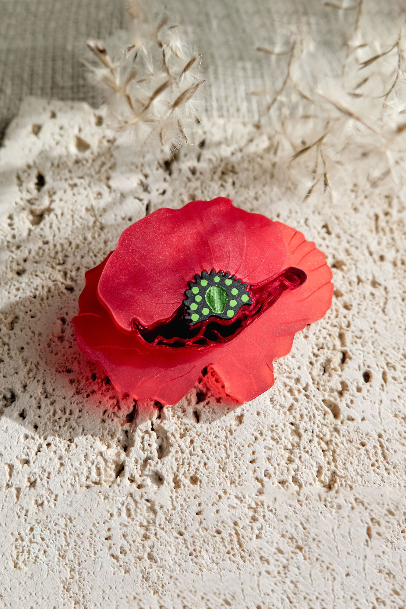 A red brooch clip shaped like a flower with a green acrylic glass center rests on a textured, off-white stone surface, accented by delicate dried plants in the background.