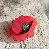 A decorative red poppy-shaped brooch clip with a green center rests on a textured cream-colored stone surface, with delicate dried plant stems in the background.