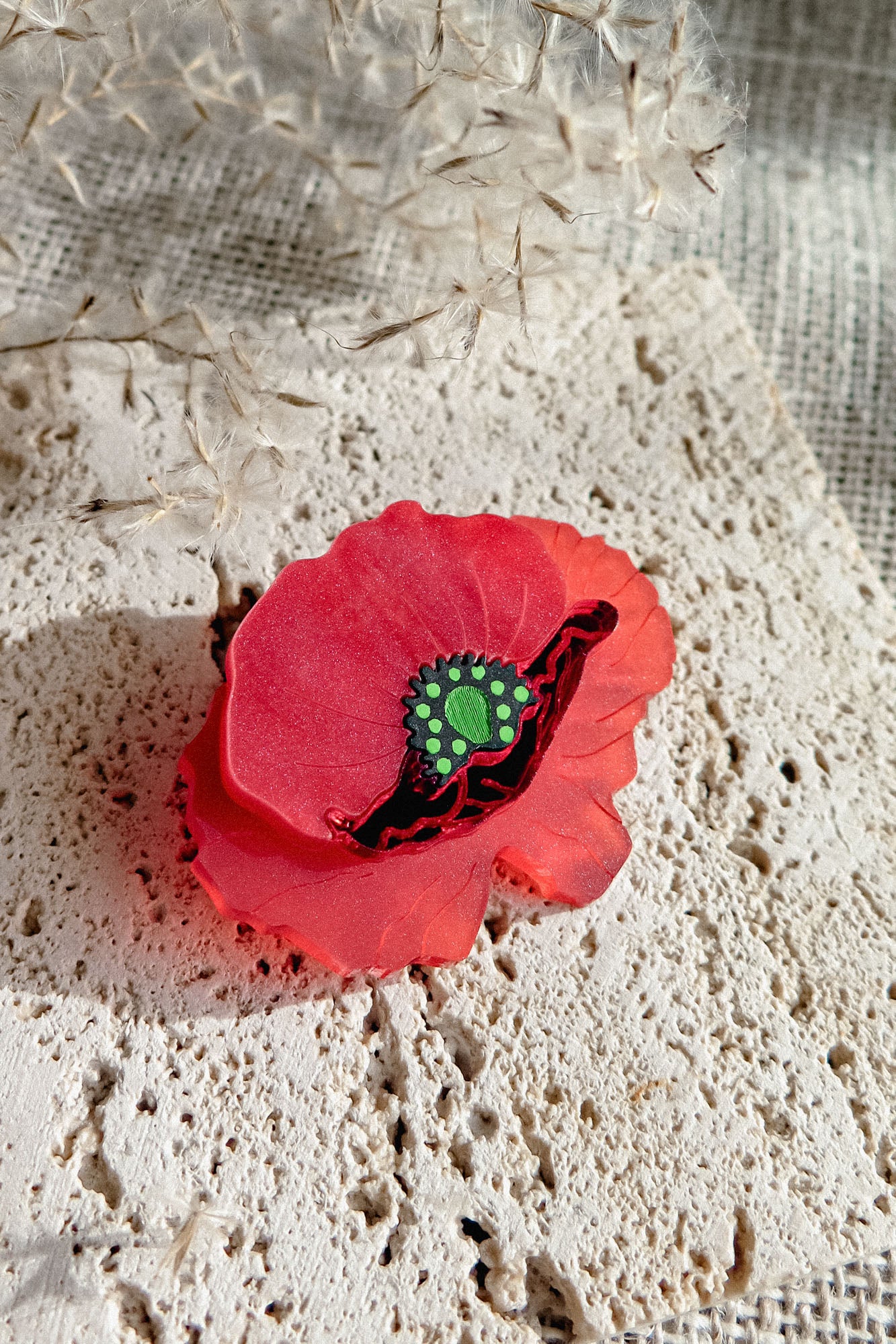 A decorative red poppy-shaped brooch clip with a green center rests on a textured cream-colored stone surface, with delicate dried plant stems in the background.