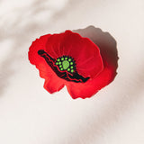 A single red poppy flower brooch with a green and black center lies on a light surface, casting a soft shadow. Subtle patterns of light and shadow appear in the background, highlighting the acrylic glass details of this elegant accessory.