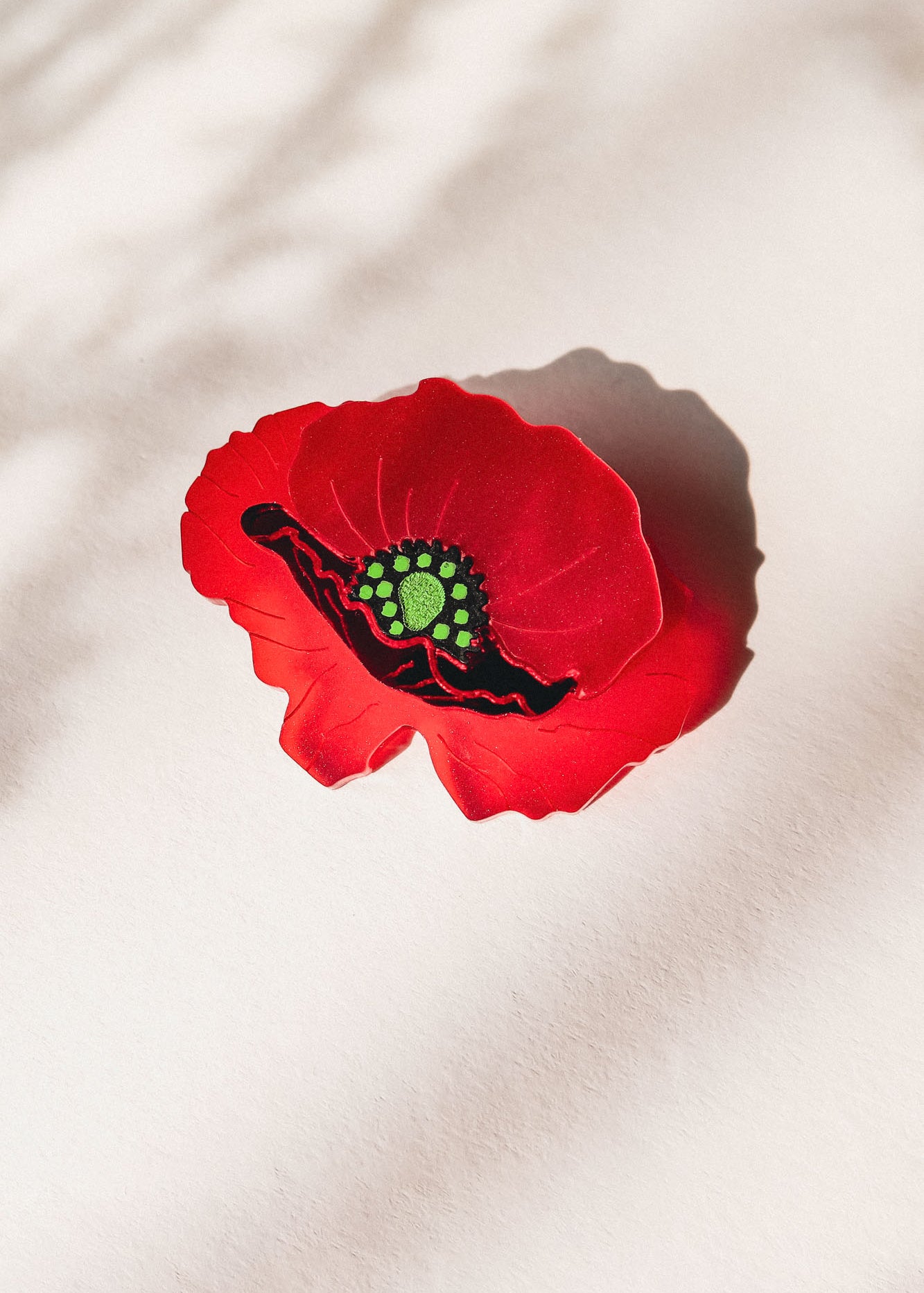 A single red poppy flower brooch with a green and black center lies on a light surface, casting a soft shadow. Subtle patterns of light and shadow appear in the background, highlighting the acrylic glass details of this elegant accessory.