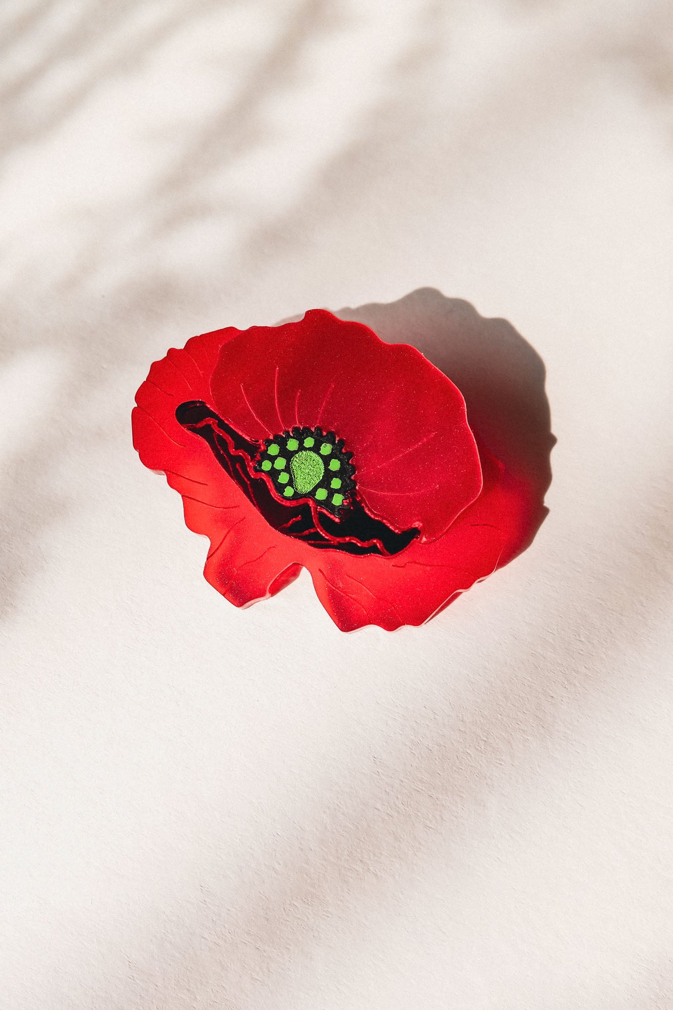 A single red poppy flower brooch with a green and black center lies on a light surface, casting a soft shadow. Subtle patterns of light and shadow appear in the background, highlighting the acrylic glass details of this elegant accessory.