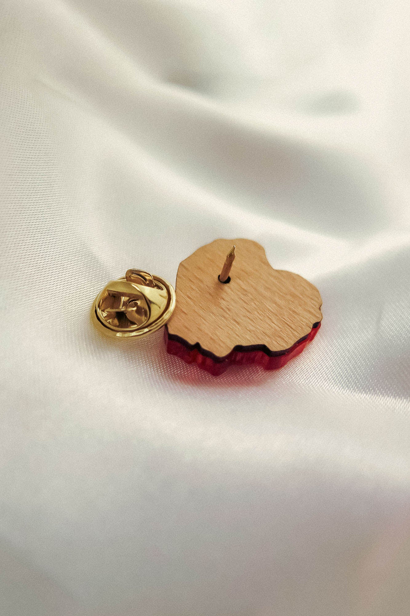 A handmade wooden pin shaped like a heart with a gold butterfly clutch backing rests on white satin fabric. Perfect for fashion enthusiasts, this piece features a red edge reminiscent of a Red Poppy Flower Lapel Pin.