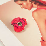 A Red Poppy Flower Lapel Pin brooch rests on an open magazine page featuring a woman with slicked-back hair in a pink top, set against a soft pink background—perfect for fashion enthusiasts seeking a chic, handmade touch.