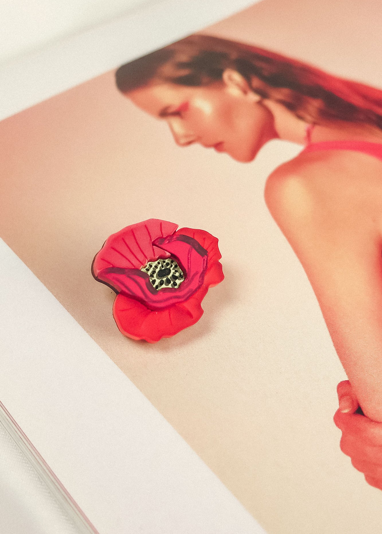 A Red Poppy Flower Lapel Pin brooch rests on an open magazine page featuring a woman with slicked-back hair in a pink top, set against a soft pink background—perfect for fashion enthusiasts seeking a chic, handmade touch.