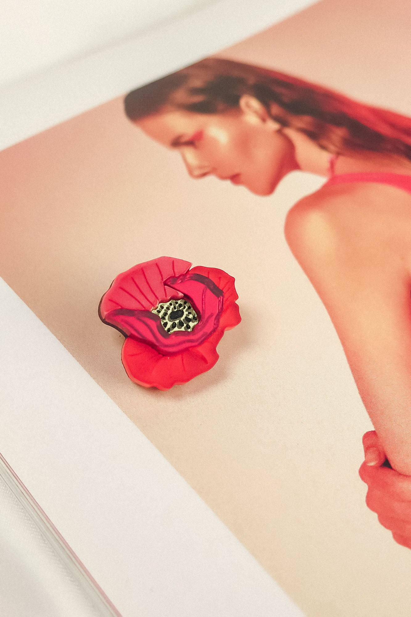 A Red Poppy Flower Lapel Pin brooch rests on an open magazine page featuring a woman with slicked-back hair in a pink top, set against a soft pink background—perfect for fashion enthusiasts seeking a chic, handmade touch.