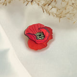 A handmade Red Poppy Flower Lapel Pin rests on a white fabric surface, with delicate dried grass or flowers in the background—an elegant accessory for fashion enthusiasts.