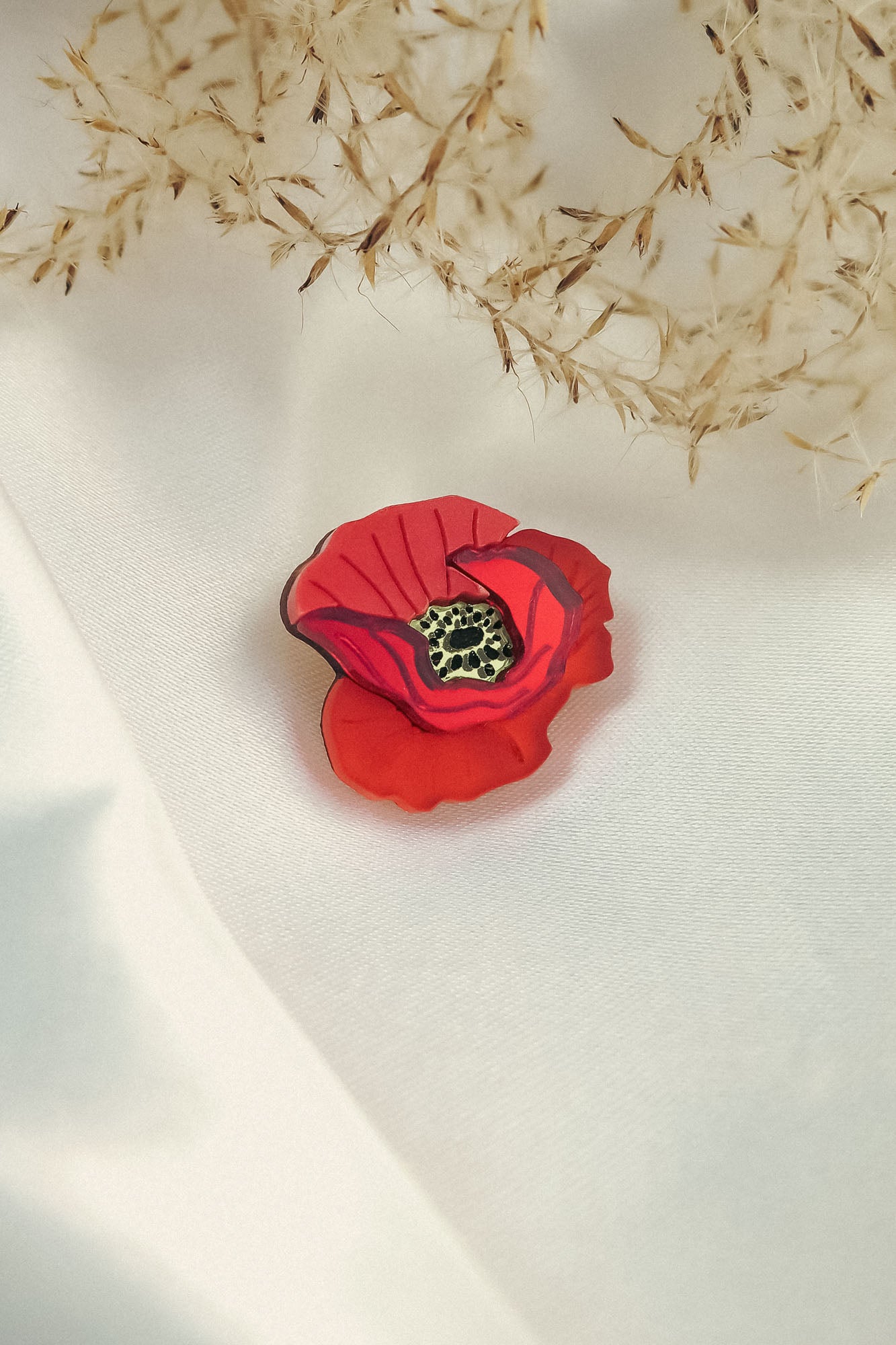 A handmade Red Poppy Flower Lapel Pin rests on a white fabric surface, with delicate dried grass or flowers in the background—an elegant accessory for fashion enthusiasts.