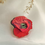 A handmade Red Poppy Flower Lapel Pin with a detailed textured center rests on soft white fabric, while delicate dried grass hangs above—an elegant accessory perfect for fashion enthusiasts.