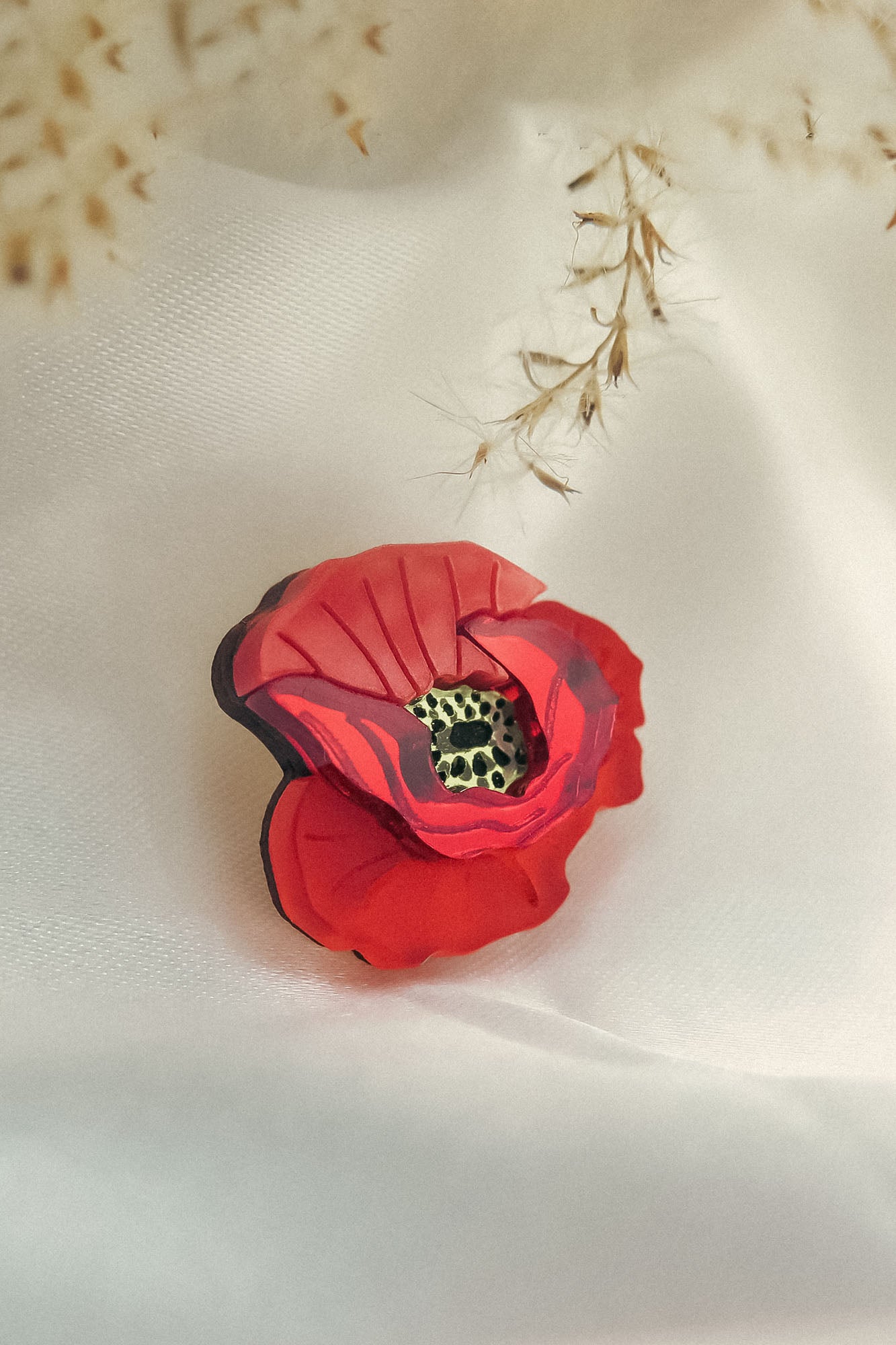 A handmade Red Poppy Flower Lapel Pin with a detailed textured center rests on soft white fabric, while delicate dried grass hangs above—an elegant accessory perfect for fashion enthusiasts.