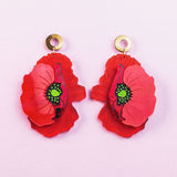 A pair of Red Poppy Statement Earrings with gold circular studs and green centers, displayed on a light pink background. This bold accessory adds vibrant flair to any look.