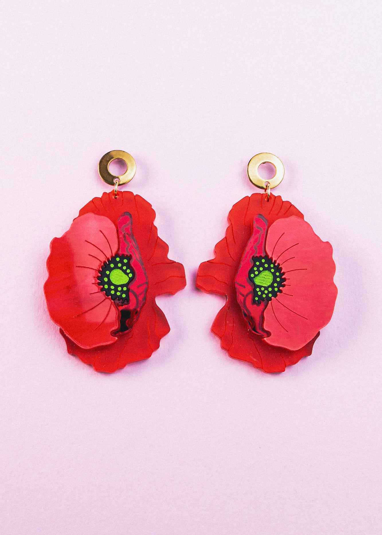 A pair of Red Poppy Statement Earrings with gold circular studs and green centers, displayed on a light pink background. This bold accessory adds vibrant flair to any look.