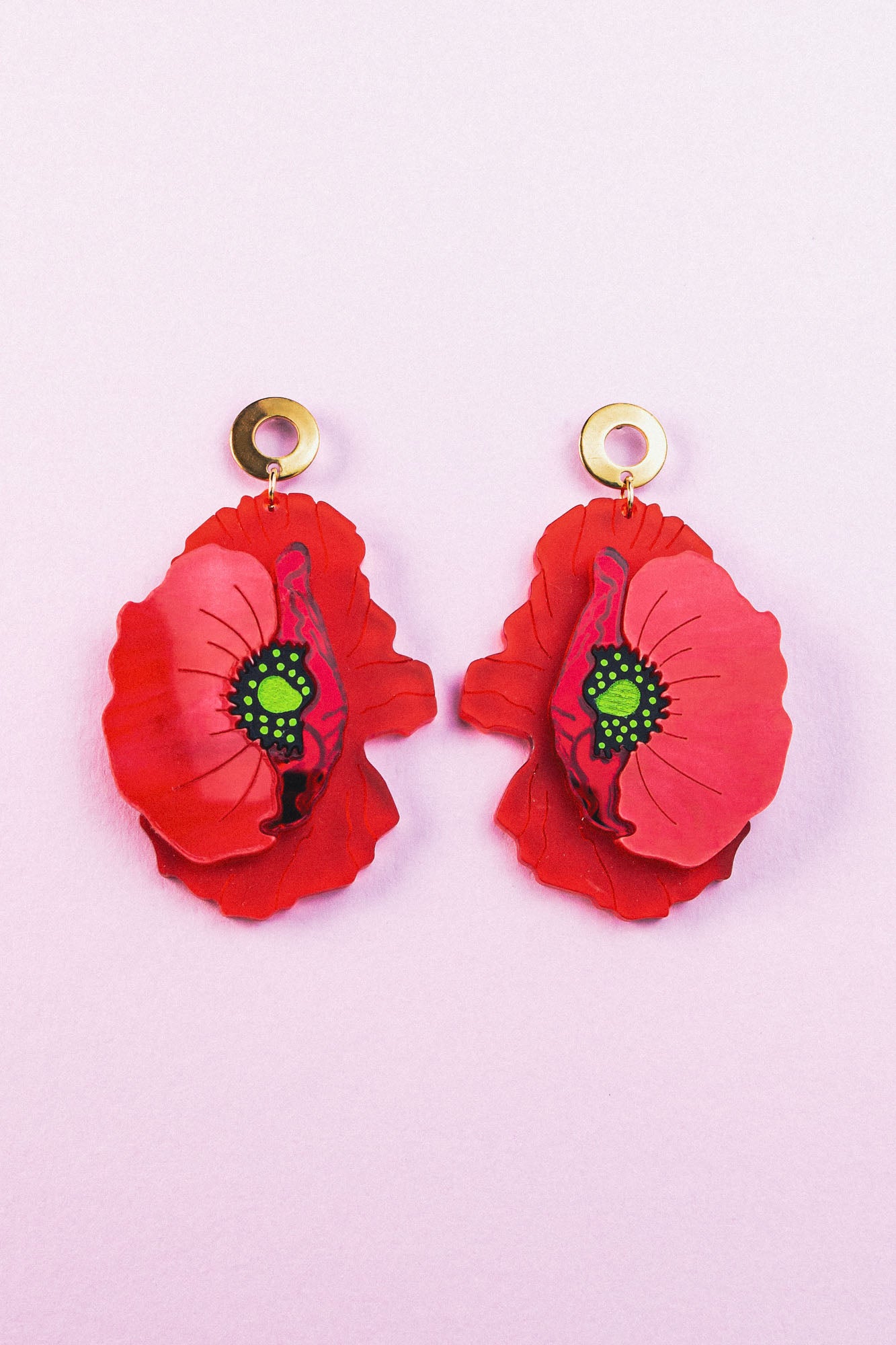 A pair of Red Poppy Statement Earrings with gold circular studs and green centers, displayed on a light pink background. This bold accessory adds vibrant flair to any look.