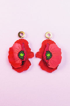 A pair of Red Poppy Statement Earrings with gold circular studs and green centers, displayed on a light pink background. This bold accessory adds vibrant flair to any look.