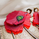 Close-up of a handcrafted Red Poppy Statement Earring with vibrant red layers and a green-black center, set against textured gold and white fabric. This bold accessory features a gold circular attachment at the top.