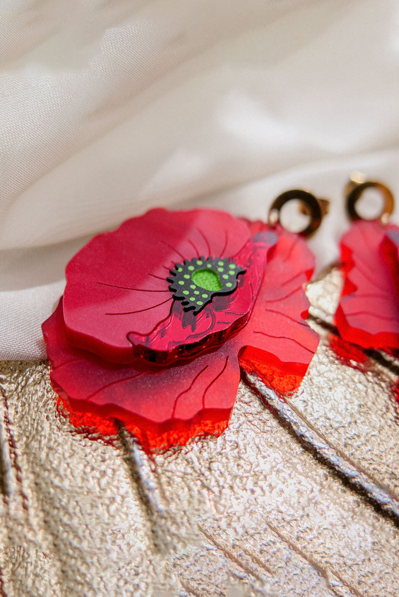 Close-up of a handcrafted Red Poppy Statement Earring with vibrant red layers and a green-black center, set against textured gold and white fabric. This bold accessory features a gold circular attachment at the top.