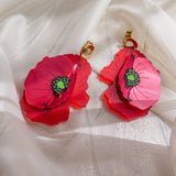 A pair of handcrafted Red Poppy Statement Earrings featuring vibrant red flower shapes with green centers and gold hooks, displayed on a white, slightly shimmery fabric—an unmistakably bold accessory.