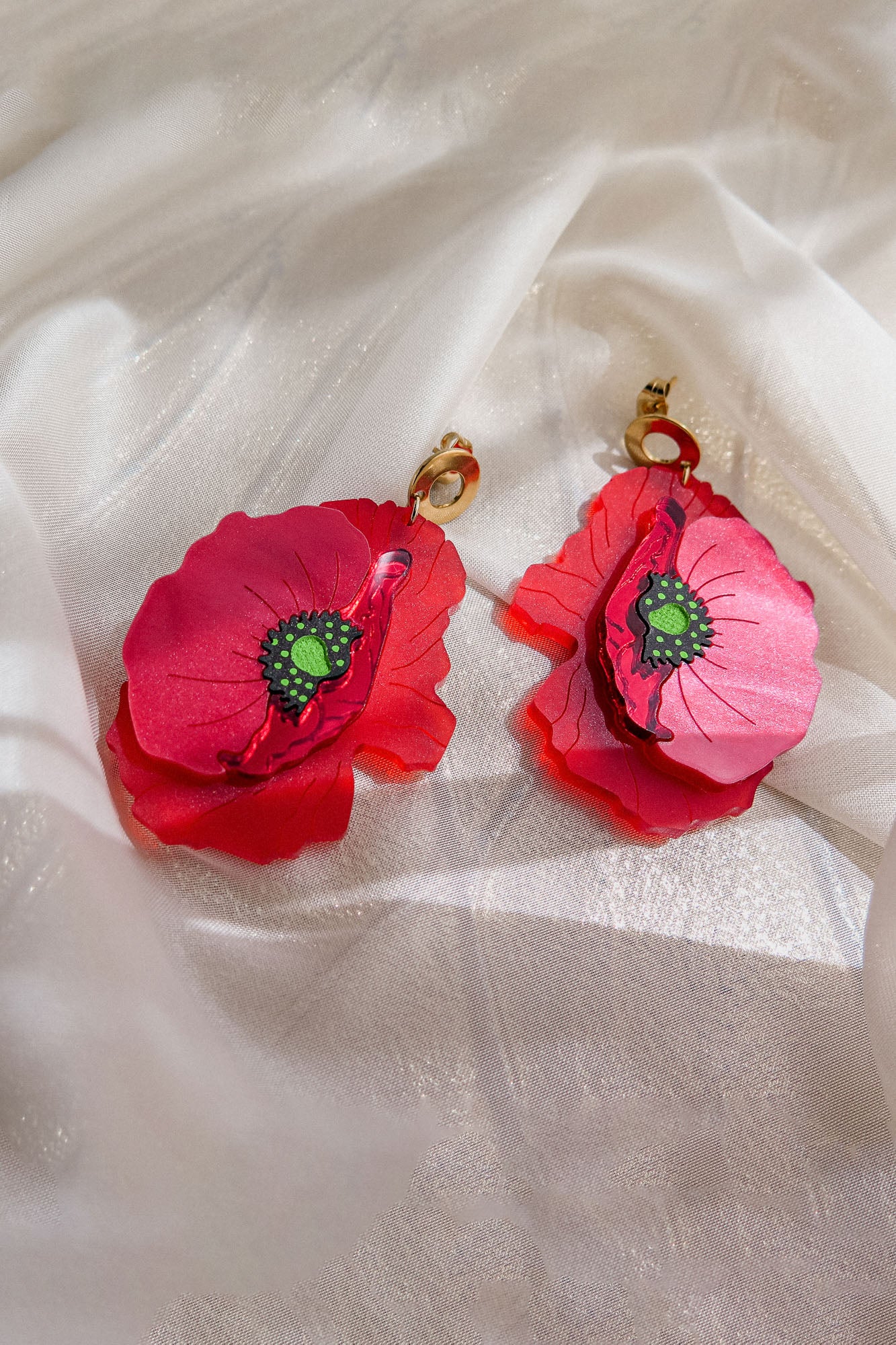 A pair of handcrafted Red Poppy Statement Earrings featuring vibrant red flower shapes with green centers and gold hooks, displayed on a white, slightly shimmery fabric—an unmistakably bold accessory.