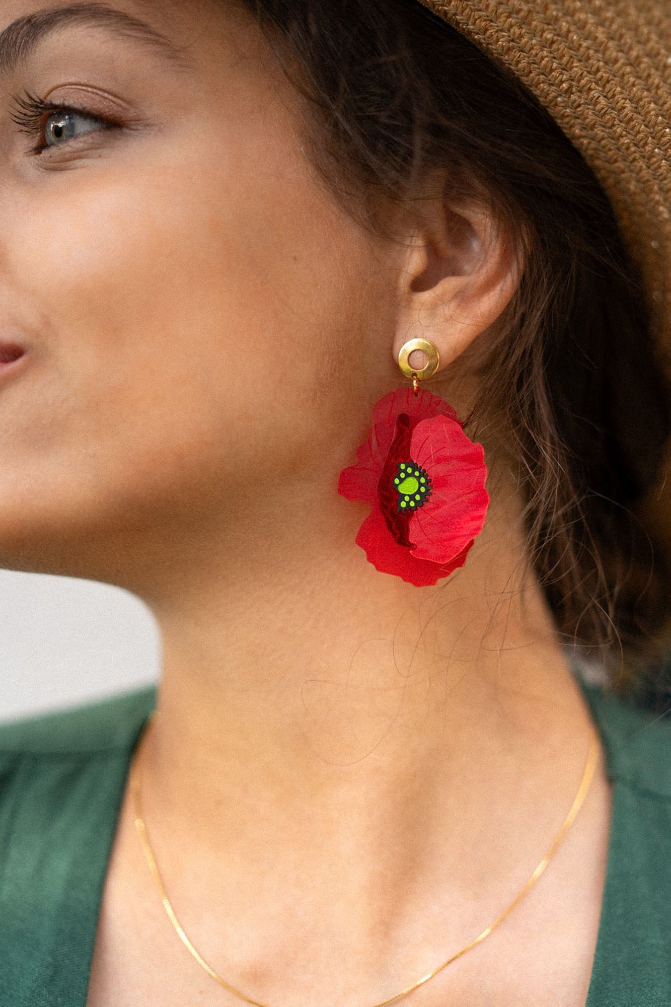 Close-up of a woman in a wide-brimmed hat, green top, and delicate gold necklace, wearing handcrafted Red Poppy Statement Earrings—a bold accessory with a vibrant red flower shape and green and black center. Her hair is tied back, and she smiles slightly.