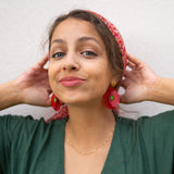 A woman with light brown hair wearing a red headscarf, handcrafted Red Poppy Statement Earrings, and a green V-neck dress stands against a light-colored wall, smiling slightly with her hands touching her head.