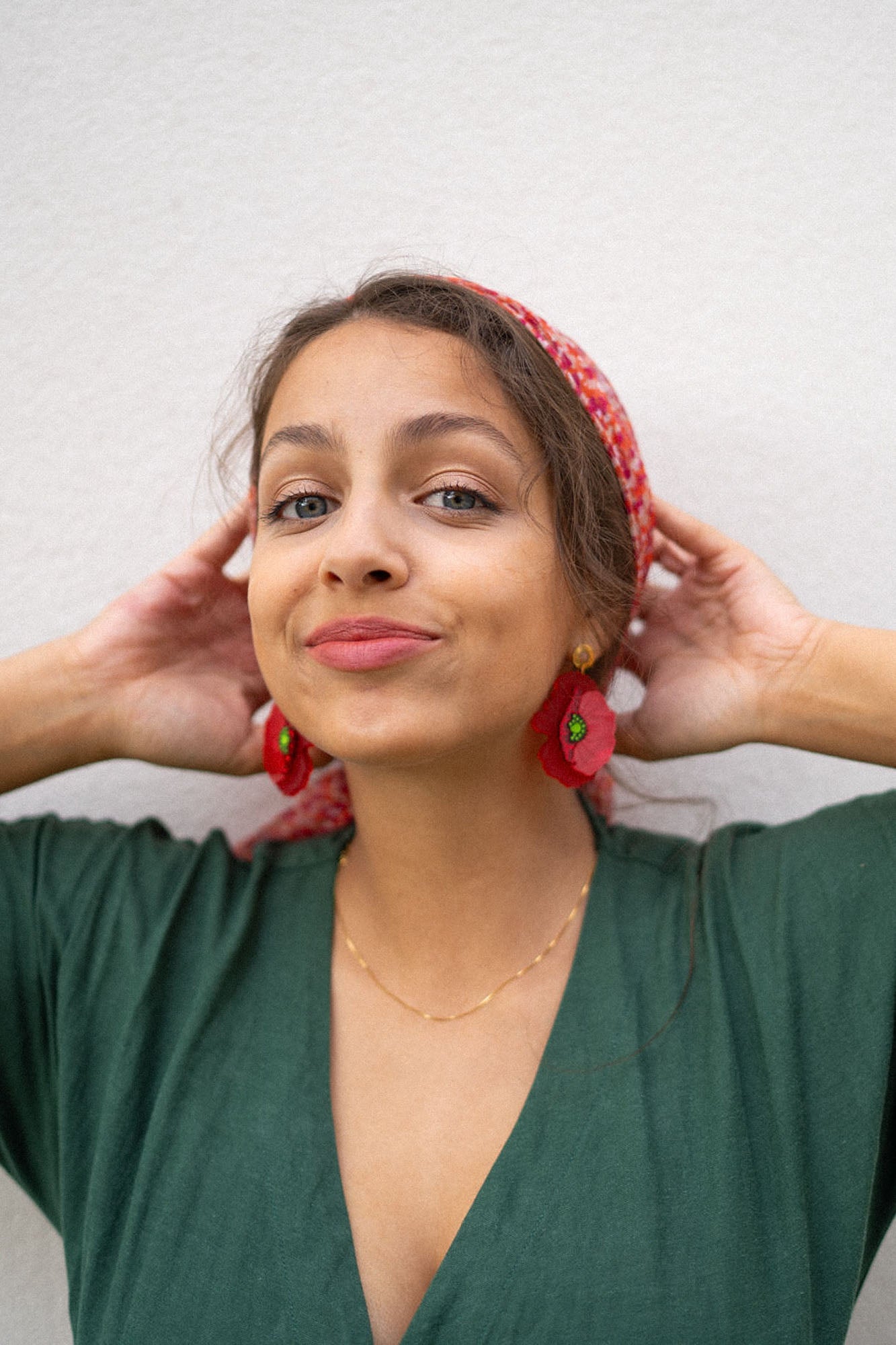 A woman with light brown hair wearing a red headscarf, handcrafted Red Poppy Statement Earrings, and a green V-neck dress stands against a light-colored wall, smiling slightly with her hands touching her head.