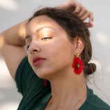 A woman with light skin and brown hair pulled back poses with one arm raised. She wears a green top, handcrafted Red Poppy Statement Earrings, and a delicate gold necklace, as sunlight and shadows fall across her face.