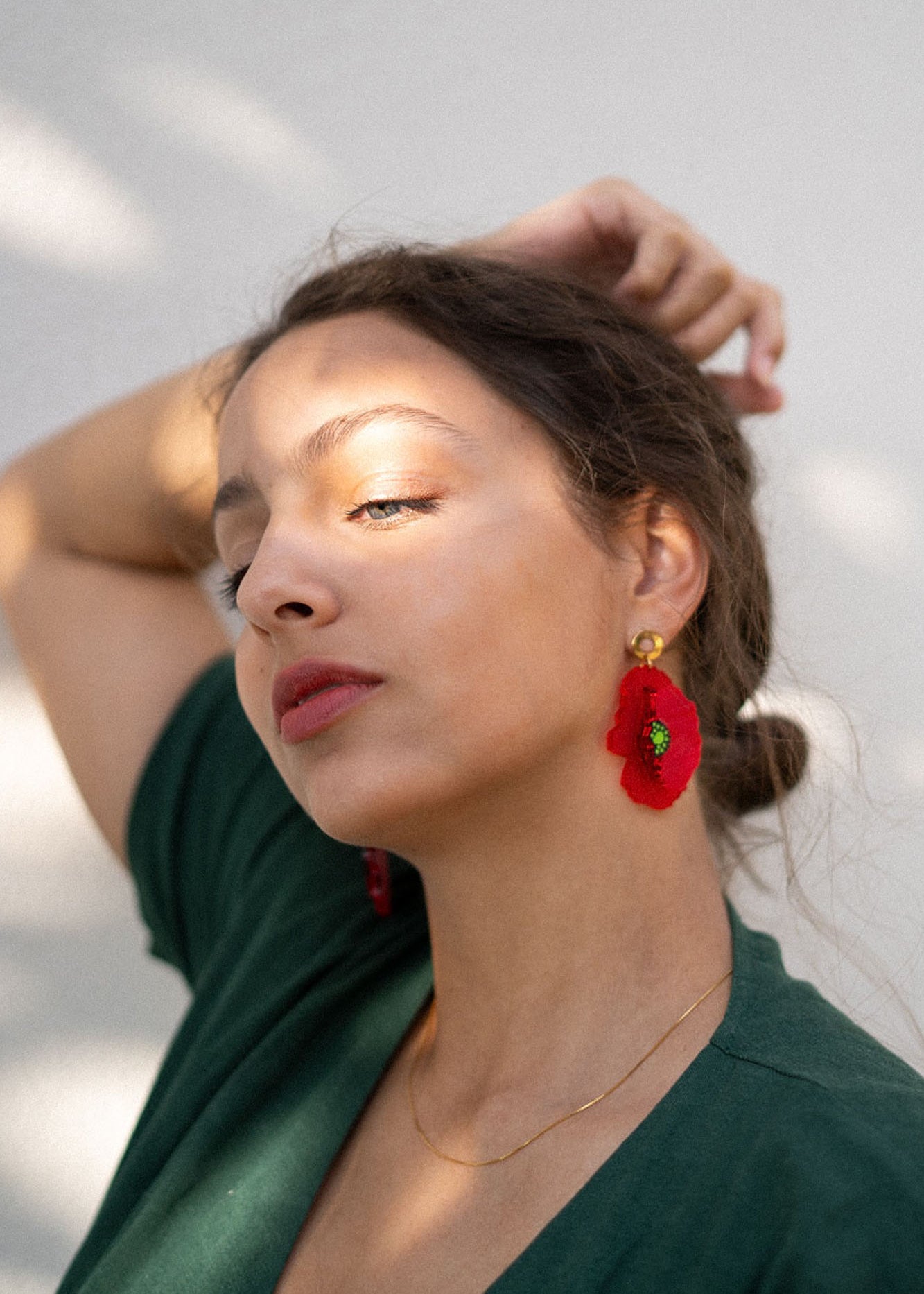 A woman with light skin and brown hair pulled back poses with one arm raised. She wears a green top, handcrafted Red Poppy Statement Earrings, and a delicate gold necklace, as sunlight and shadows fall across her face.