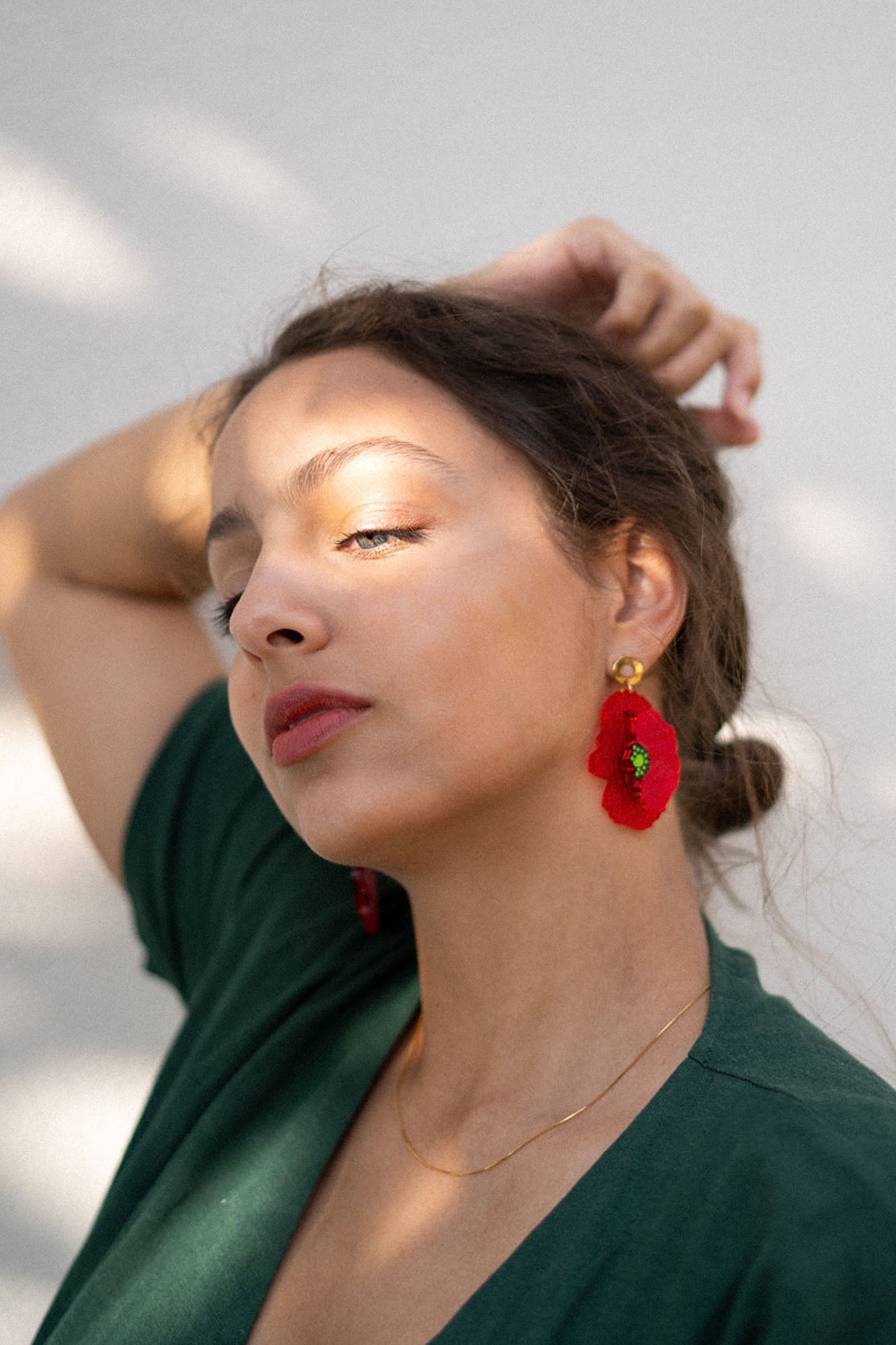 A woman with light skin and brown hair pulled back poses with one arm raised. She wears a green top, handcrafted Red Poppy Statement Earrings, and a delicate gold necklace, as sunlight and shadows fall across her face.