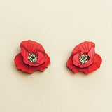 Two red poppy design acrylic earrings with black and white centers are displayed on a neutral beige background, showcasing handcrafted detail.